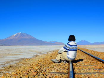 Rear view of woman sitting on railroad track against mountains and sky