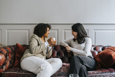 Excited female entrepreneurs having coffee and sitting on couch at work place