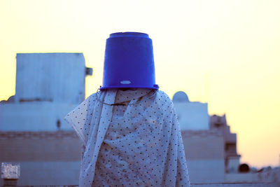 Woman with bucket on head and wrapped in textile standing against sky during sunset