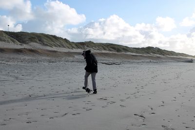 Rear view of mid adult man playing with dog while walking at beach against sky