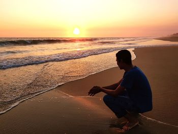 Side view of man crouching on beach against sky during sunset