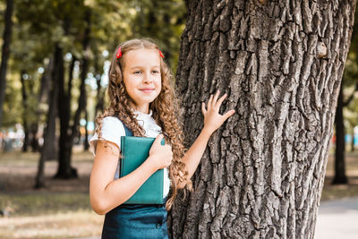 A cheerful female student in a school uniform with a book stands near a tree in the park 
