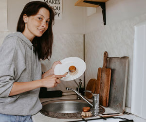 Portrait of young woman standing against wall and washing dishes