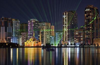 Illuminated buildings by river against sky at night