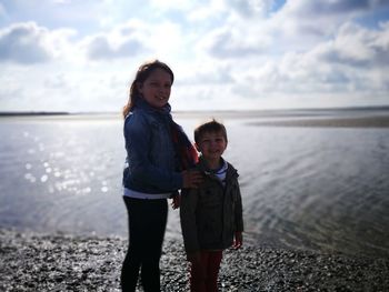 Portrait of smiling siblings standing at beach against sky during sunny day