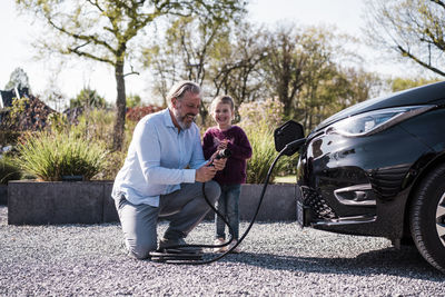 Happy man holding electric plug by daughter in front of car on sunny day