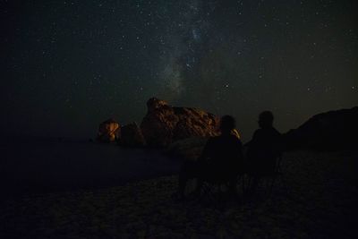 Rear view of people sitting on rock against sky at night