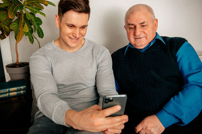 Smiling man using phone sitting with grandfather at home