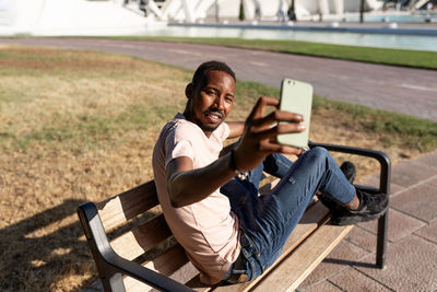 Side view of relaxed african american male sitting on bench in urban park and taking a selfie with his phone