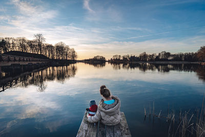 Rear view of mother and son on lake against sky during sunset