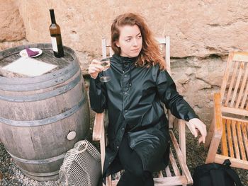 Thoughtful young woman sitting with wine on chair by wall