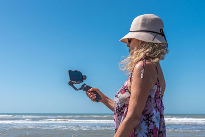 Blonde woman with a hat taking a selfie on the beach using a mobile attached to a gimbal