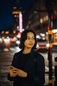 Young woman looking away while using phone in city at night