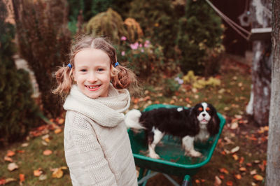 Portrait of smiling girl standing by dog in wheelbarrow on field