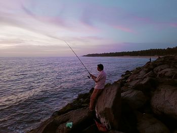 Rear view of man standing on rock by sea against sky during sunset