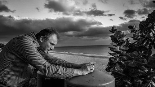 Side view of man photographing plant at beach against sky