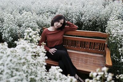 Portrait of woman sitting on bench amidst flowering plant
