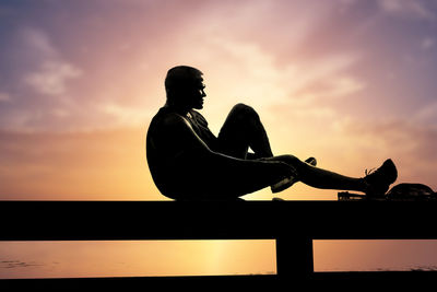 Silhouette man sitting on railing against sky during sunset
