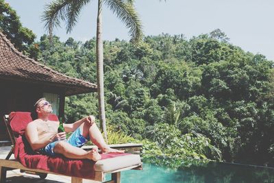 Man relaxing on lounge chair by swimming pool against trees