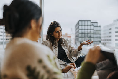 Young businesswoman gesturing while sitting by colleagues during conference meeting