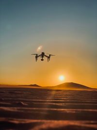 Low angle view of silhouette drone flying over land against sky during sunset