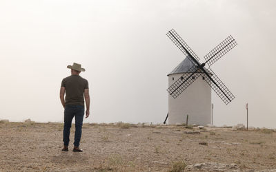 Rear view of man with cowboy hat standing on field with spanish white windmills against sky