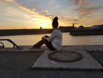 Rear view of woman sitting on retaining wall against sky during sunset