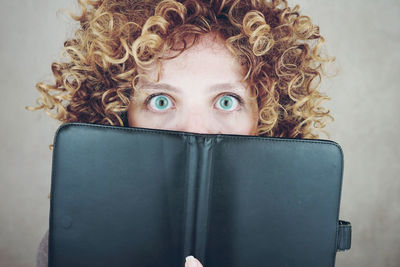 Close-up portrait of shocked young woman covering mouth with book against gray background