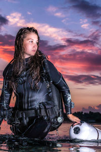 Young woman carrying diving equipment in sea against orange sky during sunset