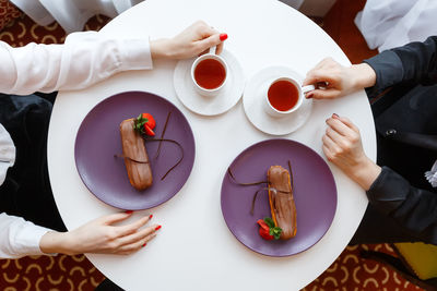 Hands of two women sitting at a table with purple plates with chocolate eclairs and two cups of tea.