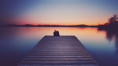 Man sitting on pier over lake against sky during sunset