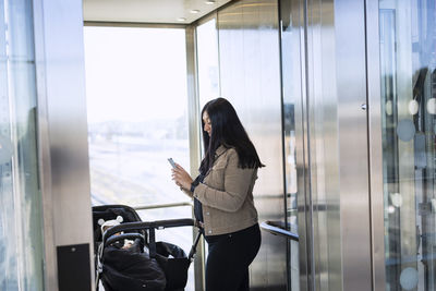 Smiling woman on parental leave with pram using cell phone inside lift