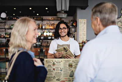Smiling owner looking at mature couple with menu in restaurant