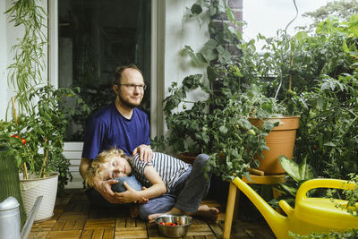 Son relaxing on father's lap while sitting amidst tomato plants in balcony