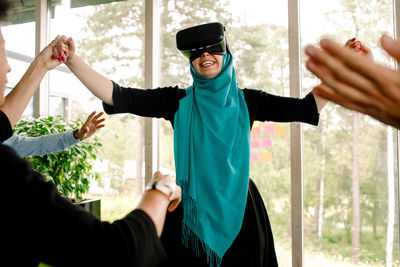 Cheerful businesswoman holding hand of colleague while using virtual reality simulator during kick off meeting at conven