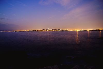 Illuminated city by sea against sky at sunset