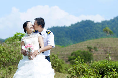 Happy bridegroom kissing bride on cheek against mountains