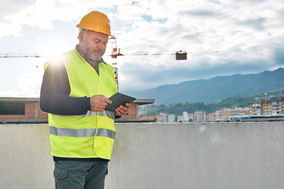 Portrait of middle aged bearded supervisor in hardhat and safety vest with tablet on building site.