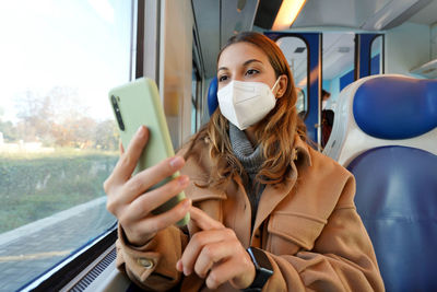 Woman traveling on train wearing medical protective face mask using smartphone. low angle.