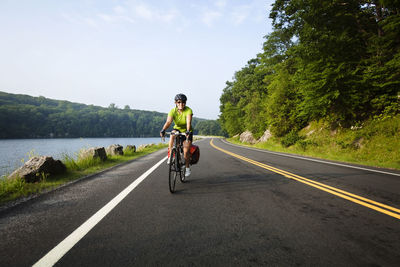 Woman cycling on road amidst trees and lake against sky