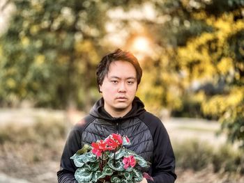 Portrait of young asian man holding red flowering cyclamen plant against trees and setting sun.
