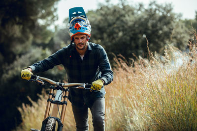 Handsome man in helmet walking looking down while carrying mountain biking bike after practice in meadow