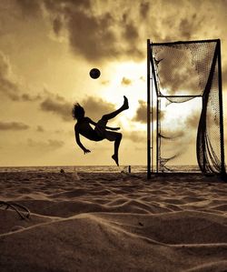 Side view of silhouette man jumping and kicking ball at beach