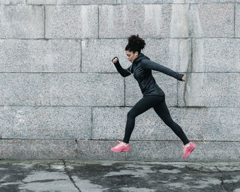 Full length of young woman in sports clothing jumping by wall