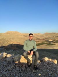 Full length of man sitting on rocks against clear blue sky