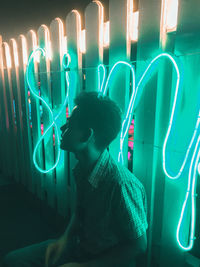 Man sitting against turquoise neon sign on fence at night