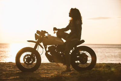 Woman sitting on motorcycle at beach during sunset