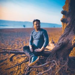 Young man sitting on land by sea against sky