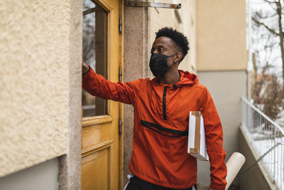 Young delivery man wearing protective face mask while standing outside house with packages