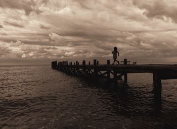 Silhouette girl running on pier over sea against cloudy sky